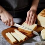 Person slicing a loaf of white bread on a wooden cutting board, with slices laid out neatly beside and a bread basket nearby.