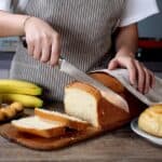 A person in an apron slices a loaf of bread on a wooden board, surrounded by bananas and pastries, with a kitchen counter in the background.
